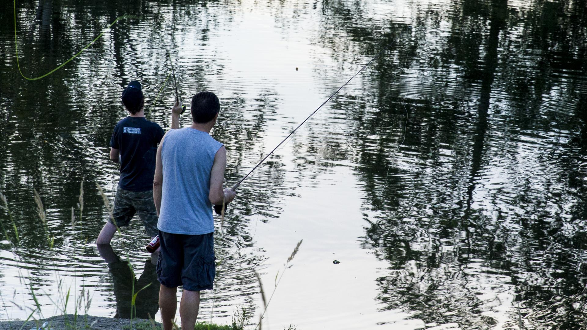 Two men stand at the edge of a lake, one standing in ankle deep water. The one on the shore holds a fishing rod. 